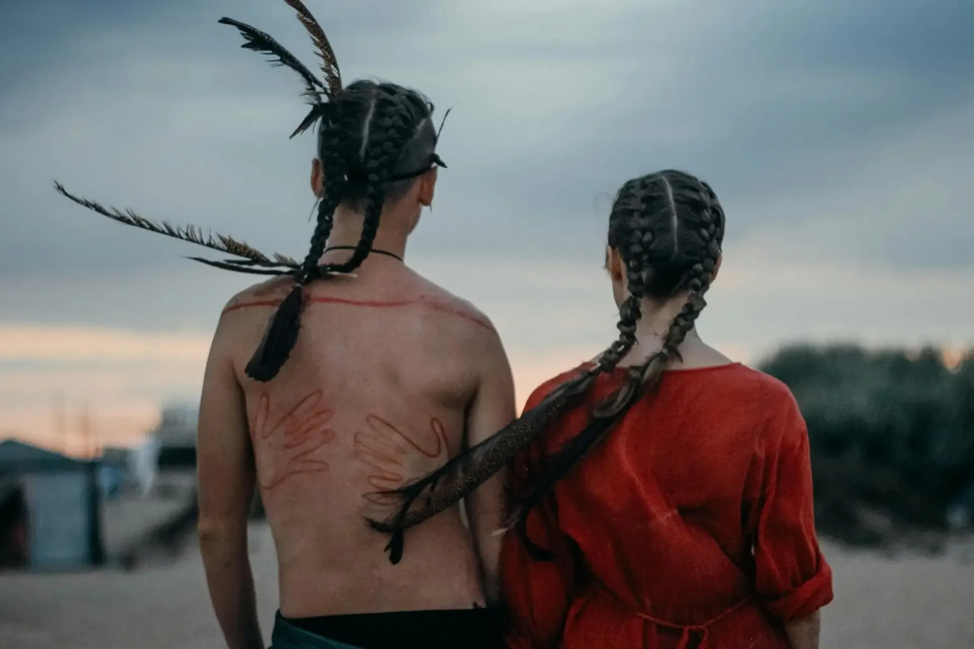 Un couple de dos, les cheveux tressés ornés de plumes, face à un paysage au crépuscule