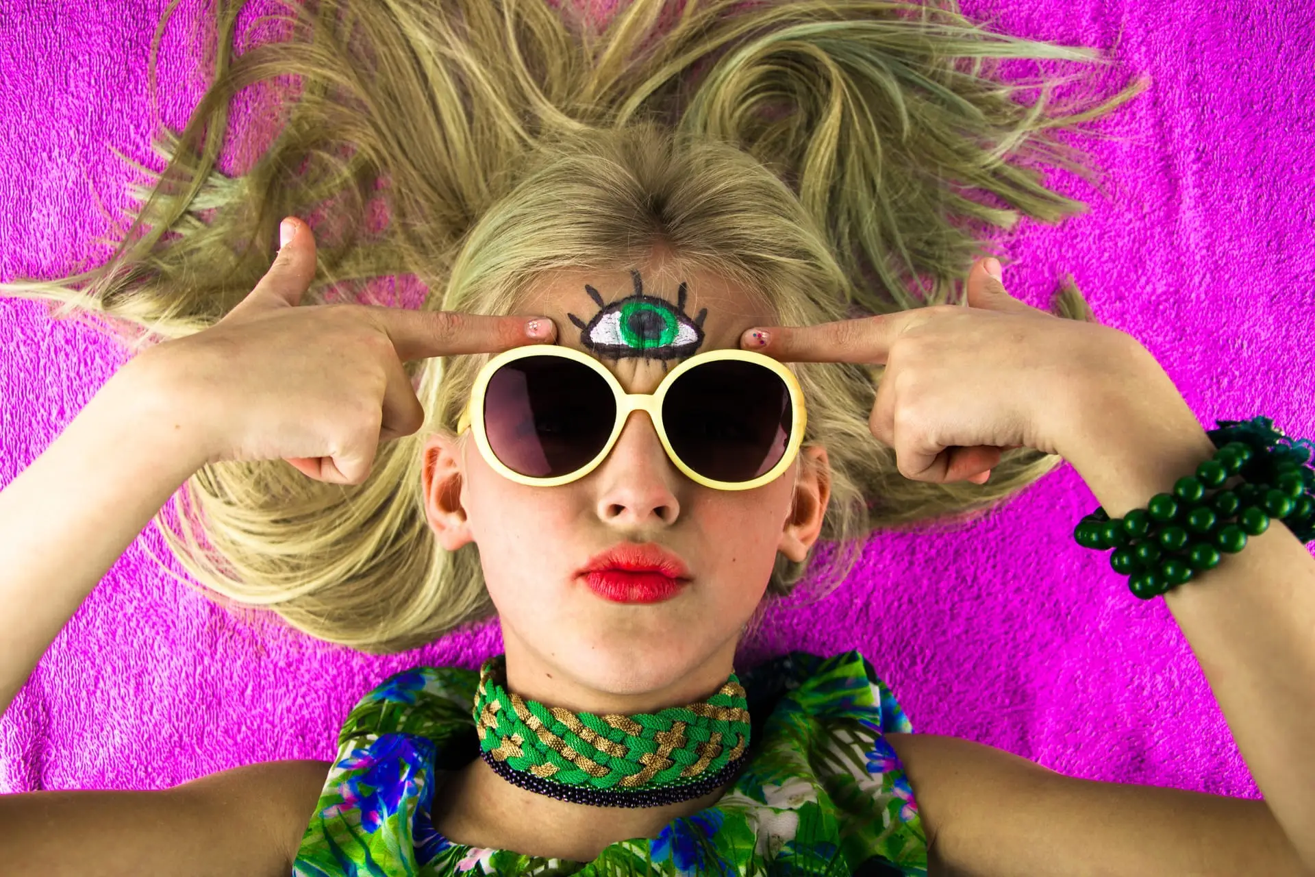 Une femme allongé avec des lunettes de soleil jaune qui pointe du doigt avec ses deux main un 3ème oeil peint sur le front