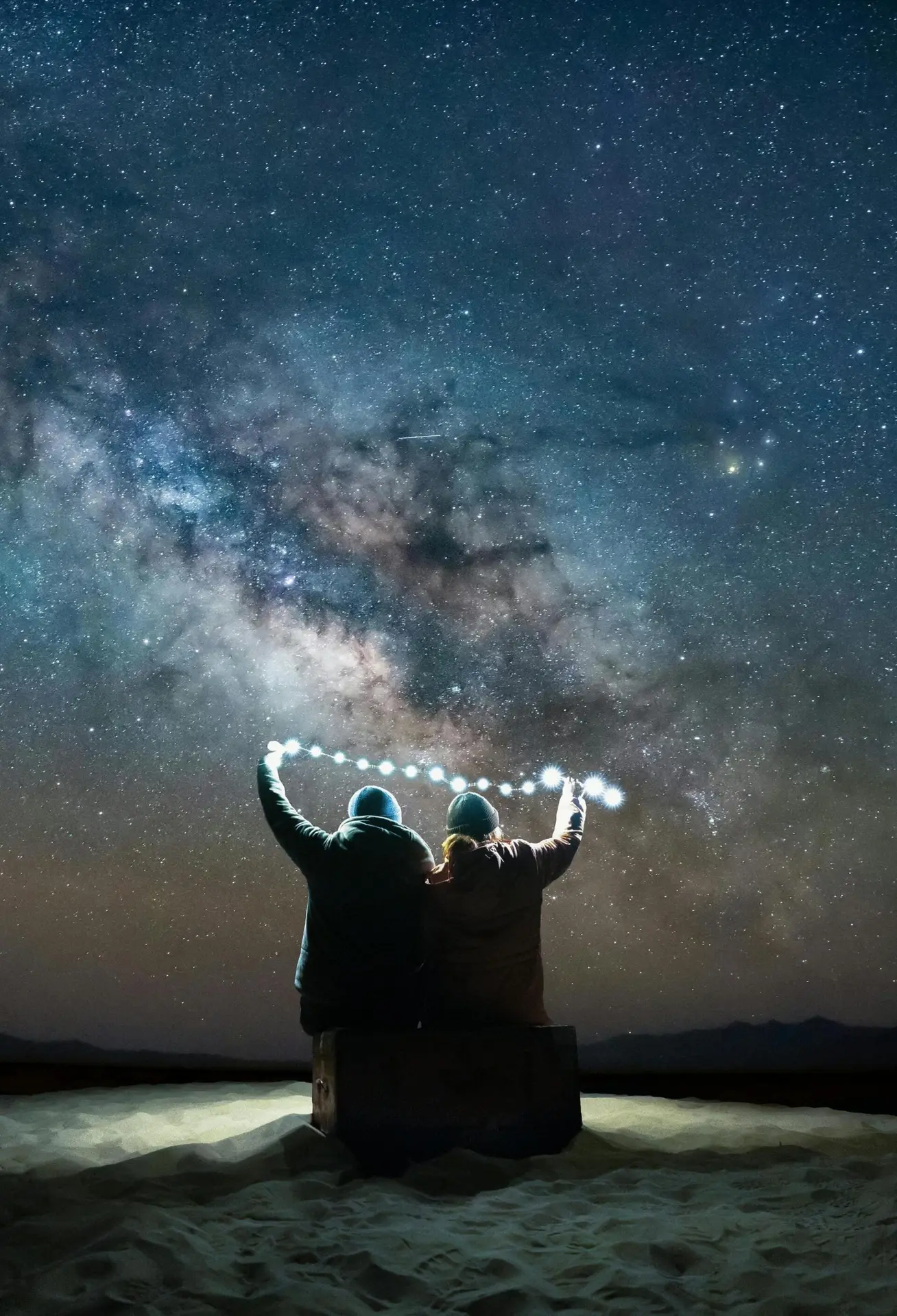 Photo d'un couple assis de dos qui tiennent une guirlande ensemble, en fon dun ciel étoilé de nuit.
